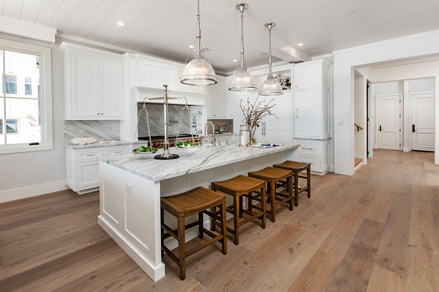 A coastal style white marble kitchen with an island with 4 brown stool and 3 glass lights overhead with a shiplap accent ceiling